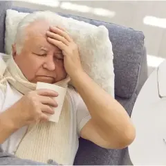 Starszy mężczyzna leży na kanapie, trzymając chusteczkę i przykładając rękę do czoła. Czuje gorączkę ze zmęczenia. Obok leżą okulary i termometr.