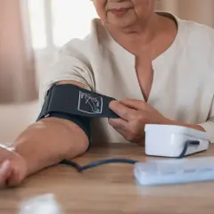 Starsza kobieta kontroluje ciśnienie w wieku 60 lat. Na stole leży ciśnieniomierz i pudełko z lekami.