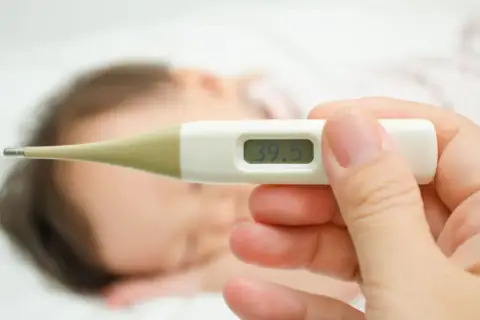 Cyfrowy termometr pokazuje 39.5°C. Dziecko śpi w tle.