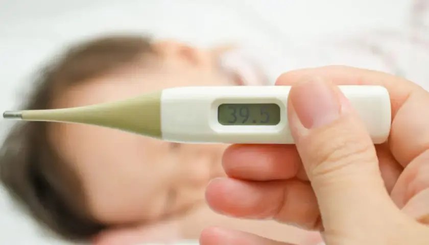 Cyfrowy termometr pokazuje 39.5°C. Dziecko śpi w tle.